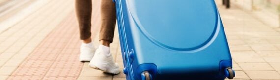 Person in white sneakers wheeling a blue hardshell suitcase along a paved walkway. Person in white sneakers wheeling a blue hardshell suitcase along a paved walkway.